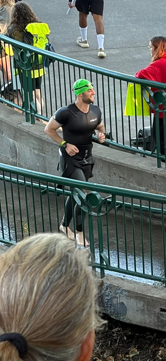 Athlete in black triathlon suit and green swim cap running during race transition, smiling while passing metal railings with spectators in background