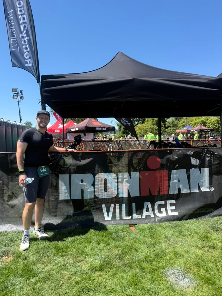 Athlete standing in triathlon gear next to Ironman Village sign under blue sky, wearing race number 644 and holding phone